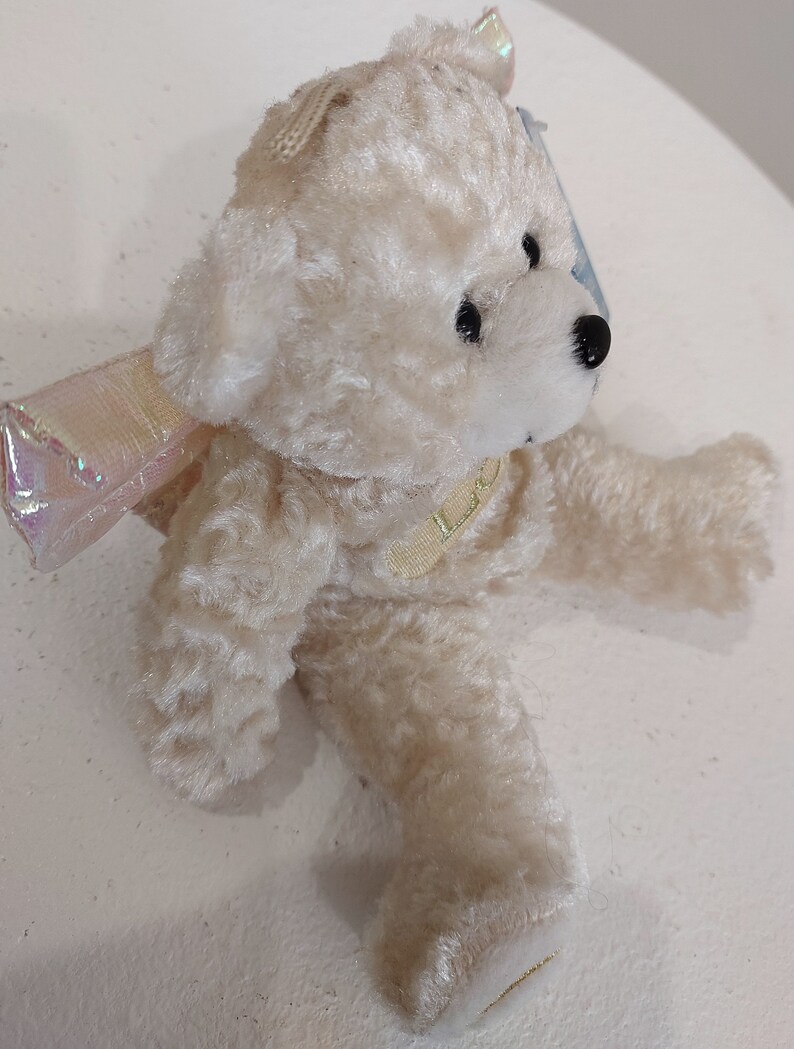 May include: A white plush teddy bear with iridescent wings. The bear is wearing a gold ribbon around its neck.
