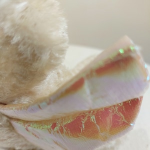 May include: A plush toy with a white fur body and iridescent pink and white wings. The wings have a shimmery, holographic finish.