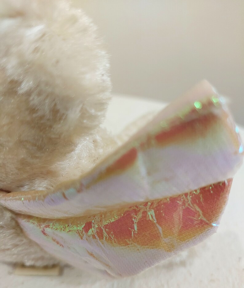 May include: A plush toy with a white fur body and iridescent pink and white wings. The wings have a shimmery, holographic finish.