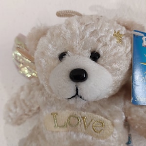 May include: A small, cream-colored teddy bear with gold wings and a gold star on its chest. The bear is wearing a gold embroidered patch that says "Love".