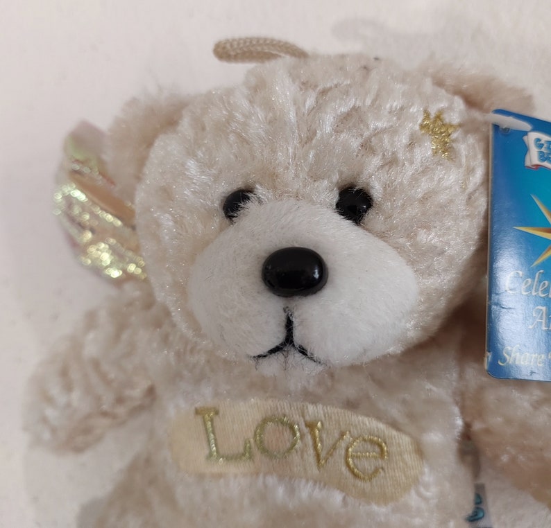 May include: A small, cream-colored teddy bear with gold wings and a gold star on its chest. The bear is wearing a gold embroidered patch that says "Love".