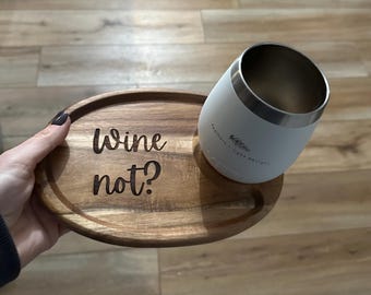May include: A white insulated wine tumbler with a silver rim rests on a wooden serving tray. The tray is engraved with the words "Wine not?" in a cursive font. The tumbler has a feather design.