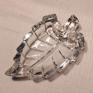 May include: A clear glass ashtray shaped like a leaf. The ashtray has a textured surface and is designed to hold cigarette butts.