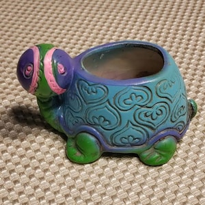 May include: A blue and green ceramic turtle planter with a pink and purple head and heart-shaped designs. The planter has a small opening at the top for planting.