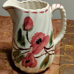 May include: White ceramic pitcher with a red, green, and brown floral design. The pitcher has a handle and a spout.