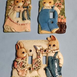 May include: Three ceramic light switch covers featuring bunny rabbits in various poses. The first cover shows a bunny in a pink dress with a blue bow, the second shows a bunny in blue overalls, and the third shows two bunnies, one in a pink dress and the other in blue overalls.