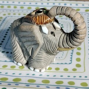 May include: A small gray ceramic elephant figurine with a brown and white patterned head and a raised trunk. The elephant has a textured surface and is decorated with small, colorful stones.