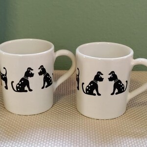 May include: Two white ceramic mugs with black handles. Each mug features a black and white illustration of a dog. The dogs are in various poses, including sitting and standing.
