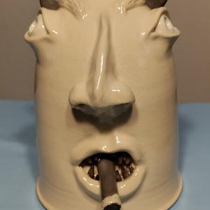 May include: A white ceramic pitcher shaped like a face with a large nose and a cigar in its mouth. The pitcher has a cork stopper and a textured surface.