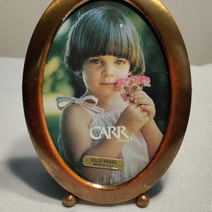 May include: Oval-shaped solid brass picture frame with a standing easel. The frame has a copper-toned finish and features a photo of a young girl holding flowers. The frame has a label that reads "Carr Framers" and "Solid Brass Made in USA."