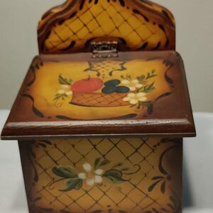 May include: A wooden box with a hinged lid, painted with a yellow and brown checkered pattern. The lid features a painted design of a basket of fruit and flowers. The box has a brown trim and a decorative handle.