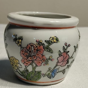 May include: A white ceramic planter with a floral design. The planter features pink, yellow, and green flowers with a red and white center. The planter has a slightly rounded shape and a wide rim.