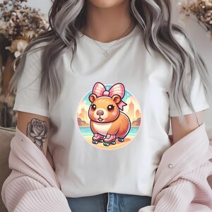 K&ouml;nnte beinhalten: Ein wei&szlig;es T-Shirt mit einem Cartoon-Bild eines braunen Hamsters, der eine rosa Schleife und Rollschuhe tr&auml;gt. Der Hamster steht vor einer Stadtkulisse mit einem Strand im Vordergrund.