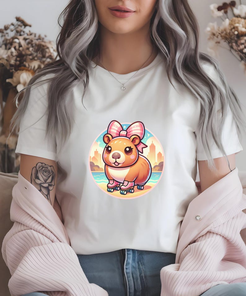 K&ouml;nnte beinhalten: Ein wei&szlig;es T-Shirt mit einem Cartoon-Bild eines braunen Hamsters, der eine rosa Schleife und Rollschuhe tr&auml;gt. Der Hamster steht vor einer Stadtkulisse mit einem Strand im Vordergrund.