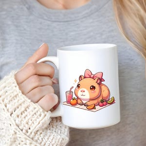 K&ouml;nnte beinhalten: Wei&szlig;e Keramiktasse mit einer Cartoon-Illustration eines Hamsters mit einer rosa Schleife, der ein Picknick mit Obst und Saft genie&szlig;t.