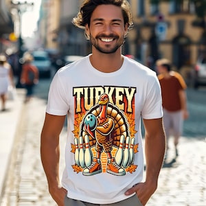 May include: White t-shirt with a graphic of a turkey holding a bowling ball, surrounded by bowling pins and autumn leaves. The word "TURKEY" is printed above the turkey. The turkey is wearing orange sneakers.