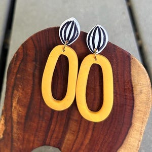 May include: A pair of statement earrings. Each earring features a bright yellow, oval-shaped pendant suspended from a smaller, white and black striped oval. The earrings are displayed on a wooden surface.