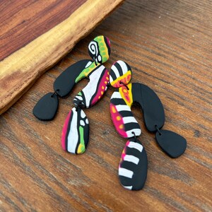 Peut inclure: Une paire de boucles d'oreilles abstraites en pâte polymère. Les boucles d'oreilles présentent une combinaison de formes géométriques noires, blanches, roses, jaunes et vertes. Les boucles d'oreilles sont sur une surface en bois.