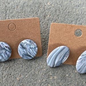 May include: Two pairs of marbled stud earrings on brown earring display cards. One pair is round, the other is oval. The earrings have a swirling pattern of gray and white. The cards have a hole at the top for hanging.