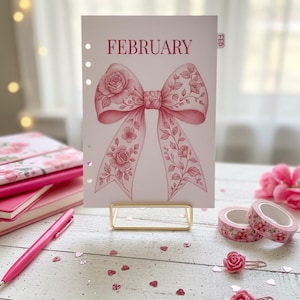 Può includere: Una pagina di pianificatore rosa con la parola "FEBRUARY" e un design a fiocco ad acquerello con accenti floreali. La pagina è tenuta in un supporto dorato, con nastro washi rosa e una penna nelle vicinanze. Il tema è floreale e romantico.