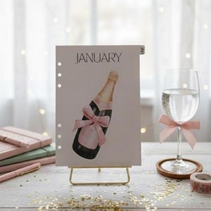 May include: A planner page for January with a champagne bottle illustration and a pink bow. A glass of water with a pink bow, a roll of floral tape, and a stack of pink notebooks are also visible. The planner is on a gold stand.