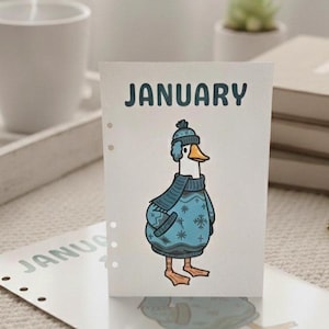 May include: A calendar page for January featuring a cartoon duck wearing a blue sweater and hat. The word "JANUARY" is printed at the top. The duck has an orange beak and feet. The calendar is on a table with a white mug and books.
