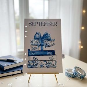 May include: A watercolor illustration of a stack of books with a blue bow, labeled "SEPTEMBER." The books have floral and patterned designs in shades of blue. The artwork is displayed on a small gold easel, with blue notebooks and decorative tape nearby.