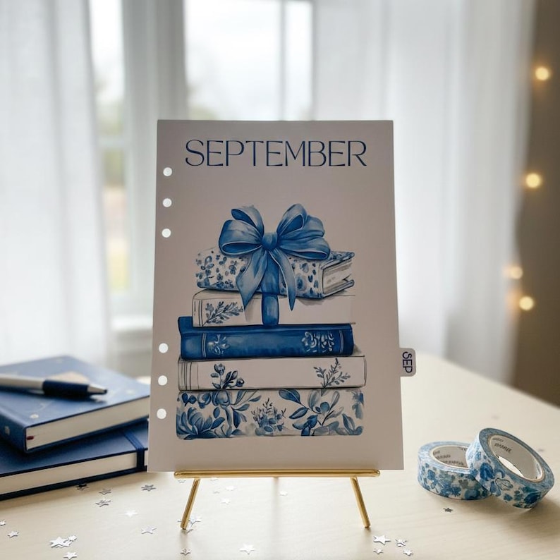 May include: A watercolor illustration of a stack of books with a blue bow, labeled "SEPTEMBER." The books have floral and patterned designs in shades of blue. The artwork is displayed on a small gold easel, with blue notebooks and decorative tape nearby.