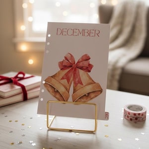 May include: A decorative calendar page for December, featuring watercolor illustrations of two golden bells with a red bow. The page is held in a gold metal stand, with a roll of patterned tape and a stack of books nearby.