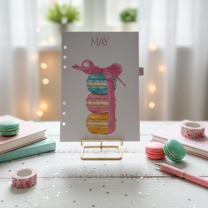 May include: A planner page with the word "MAY" at the top, featuring a watercolor illustration of stacked macarons in blue, pink, and yellow, tied with a pink ribbon. The page is held by a gold-colored stand, with other stationery items nearby.