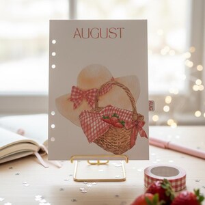 May include: A watercolor illustration of a picnic scene on a calendar page for August. The image features a straw hat with a pink bow, a picnic basket with strawberries, and the word "AUGUST" in red. The calendar is held by a gold stand.