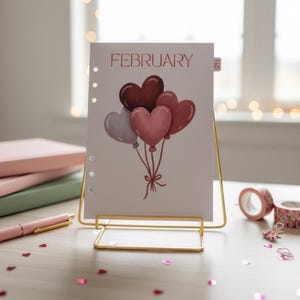 May include: A white card with the word "FEBRUARY" and heart-shaped balloons in shades of red and pink. The card is displayed in a gold metal stand, with a pink pen and notebooks in the background.