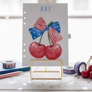 May include: A planner page for July featuring a watercolor illustration of two cherries and a bow with a red, white, and blue design. The page is held in a gold metal stand. Decorative tape and a pen are also visible.