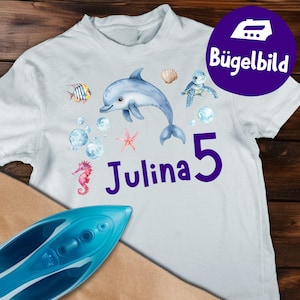 Pode incluir: Camiseta branca com um golfinho azul, criaturas marinhas, bolhas e o nome "Julina" com o número "5" em roxo. O design inclui um cavalo-marinho, uma estrela-do-mar e uma tartaruga, com o texto "Bügelbild".