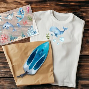 Peut inclure: Un t-shirt blanc avec une illustration &agrave; l'aquarelle d'une baleine bleue, des bulles et des algues. Une feuille d'autocollants de cr&eacute;atures marines et un fer &agrave; repasser bleu reposent sur une surface en bois. Les autocollants incluent une sir&egrave;ne, un dauphin et une pieuvre.