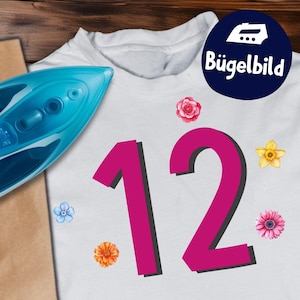 Könnte beinhalten: Weißes T-Shirt mit einer großen magentafarbenen Zahl 12 und floralen Akzenten. Ein blaues Bügeleisen und eine braune Papiertüte sind zu sehen. Ein blauer Kreis mit dem Text "Bügelbild" befindet sich oben rechts.