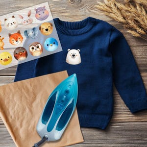 Pode incluir: Um su&eacute;ter azul marinho com um patch de urso polar branco. Uma folha de patches de animais para passar a ferro, incluindo uma raposa, uma coruja e um hipop&oacute;tamo, est&aacute; ao lado do su&eacute;ter. Um ferro azul e branco e papel castanho tamb&eacute;m est&atilde;o na imagem.