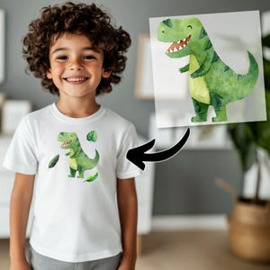 May include: White t-shirt featuring a watercolor illustration of a green dinosaur with a smiling face, red teeth, and green leaves. The dinosaur design is centered on the shirt. The image also shows a child wearing the shirt.