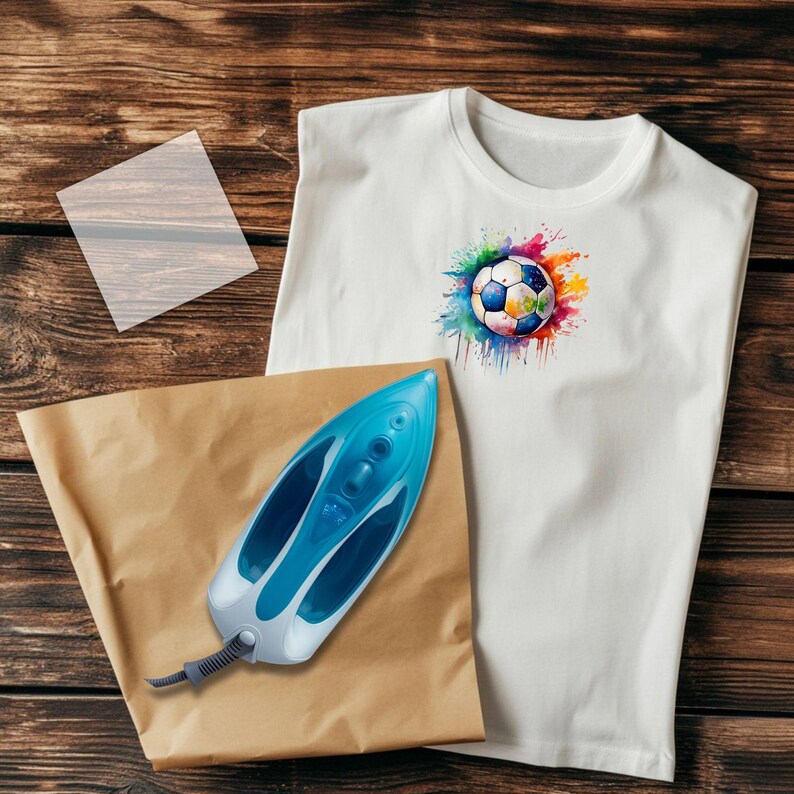 May include: White t-shirt with a colorful soccer ball graphic. A blue and white iron, a brown paper sheet, and a clear sheet are next to the shirt on a wooden surface. The soccer ball design features a watercolor effect with splashes of color.