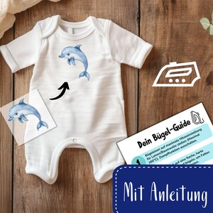 May include: A white baby onesie with a blue dolphin graphic. The onesie is shown with an iron-on transfer sheet and instructions for applying the transfer. The instructions are in German and include the text "Dein B&uuml;gel-Guide" and "Mit Anleitung".