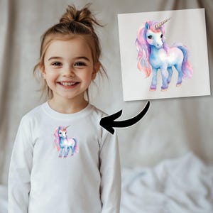 May include: A white long-sleeve shirt featuring a watercolor illustration of a unicorn with a flowing pink and purple mane and tail, and a spiraled horn. The unicorn is also depicted on a print in the background.