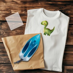 May include: White t-shirt with a watercolor illustration of a green dinosaur. A blue and white iron, a sheet of transfer paper, and a brown paper are on a wooden surface. The t-shirt is sleeveless.