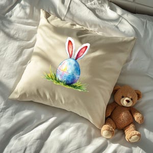 K&ouml;nnte beinhalten: Ein beigefarbener Kissenbezug mit einer Aquarell-Illustration eines Ostereis mit Hasenohren. Das Ei ist blau und wei&szlig; mit einem gelben Stern im Inneren. Die Hasenohren sind rosa und wei&szlig;. Der Kissenbezug liegt auf einem wei&szlig;en Bett mit einem braunen Teddyb&auml;r.