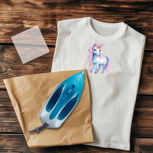 May include: White t-shirt with a watercolor unicorn design, a blue and white iron, and a brown paper sheet. The iron is approximately 12 inches long. The unicorn has a pink and purple mane and tail.