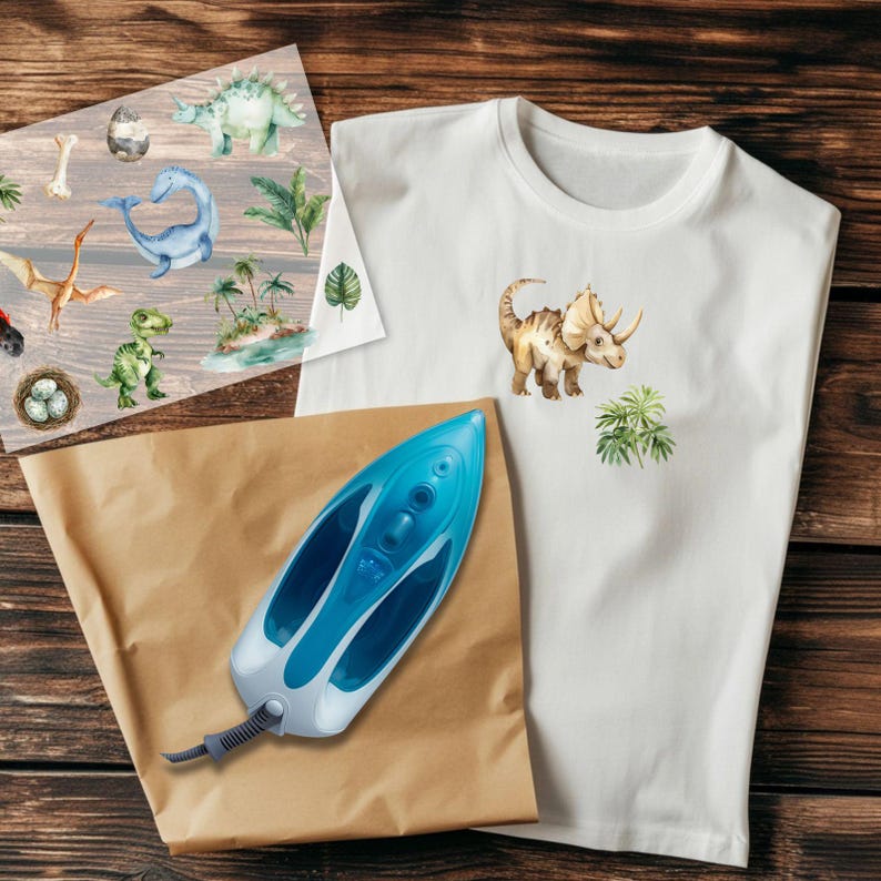 May include: A white t-shirt featuring a watercolor illustration of a triceratops and green foliage. A sheet of dinosaur-themed iron-on transfers and a blue and white iron are also present, all arranged on a wooden surface.