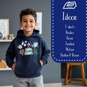 May include: A young person wearing a navy blue hoodie with a colorful underwater scene print. The print features a shark, starfish, treasure chest, coral, and fish. The text "Ideen: T-shirts, Hoodies, Hosen, Taschen, M&uuml;tzen, Decken & Kissen" is visible in the background.