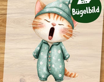 May include: A watercolor illustration of a yawning orange tabby cat wearing a teal sleep cap and pajamas with white polka dots. The cat has its mouth wide open and is standing on a light wood grain background. A green circle with the text "Bügelbild" is in the upper right corner.