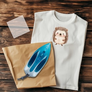 May include: A white t-shirt featuring a brown and white hedgehog illustration. A blue iron and brown paper are on a wooden surface.