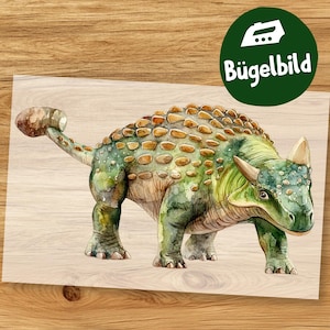 Pode incluir: Ilustração em aquarela de um dinossauro Ankylosaurus em tons de verde, marrom e bege. O dinossauro tem um corpo arredondado coberto por placas texturizadas e uma cauda em forma de clava. Um círculo verde com o texto "Bügelbild" é visível.