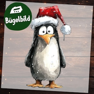 May include: A cartoon penguin wearing a red Santa hat with a white trim and pom-pom. The penguin is depicted in black, white, and grey with an orange beak and feet. The image includes the text "Bügelbild" in a green circle, suggesting an iron-on transfer.